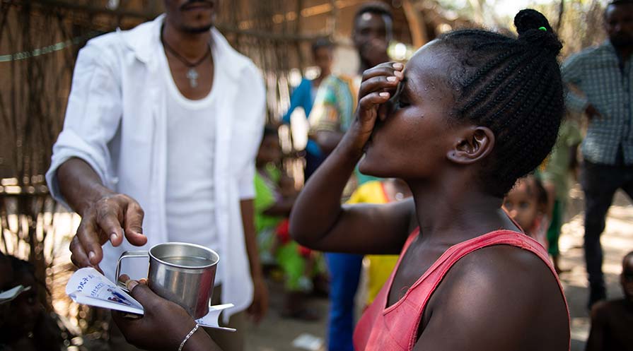 Photo of a woman in an Ethiopian village taking Mectizan with a cup of water in hand while onlookers watch in the background.