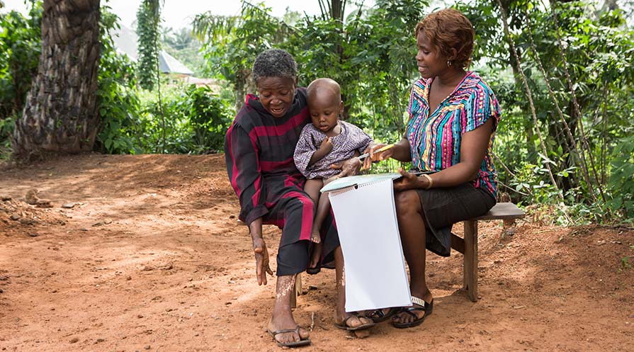 Photo of two women sitting on a wooden bench outside. One his holding a pen and paper calendar in her hand, the other is holding a young child.